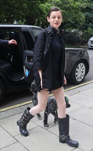 Emma Corrin wears a black short dress with black buckled boots