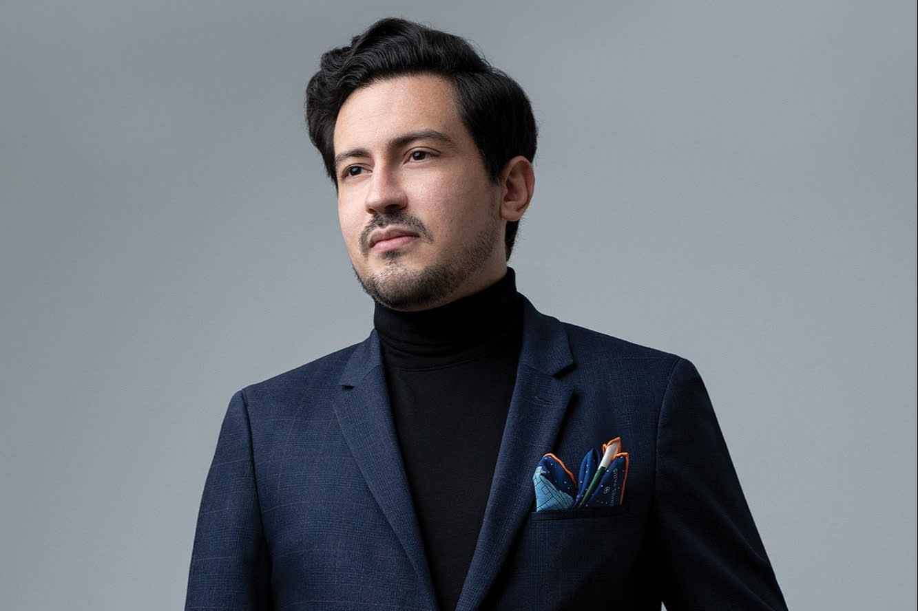 A stylishly dressed man wearing a navy blue suit, black turtleneck, and a patterned pocket square. He stands with one hand in his pocket