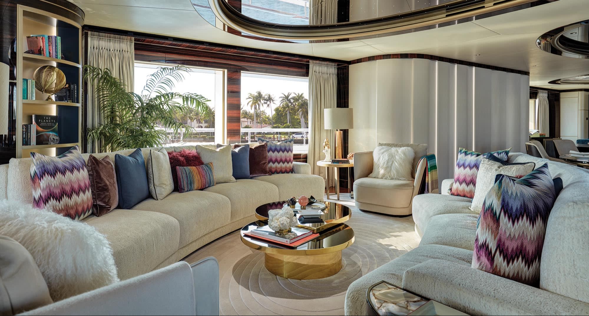 A spacious and elegantly decorated living room featuring cream-colored sofas with an array of colorful throw pillows. In the middle is a sleek gold coffee table with a reflective surface, displaying books and decorative pieces. Large windows provide natural light and a view of palm trees outside