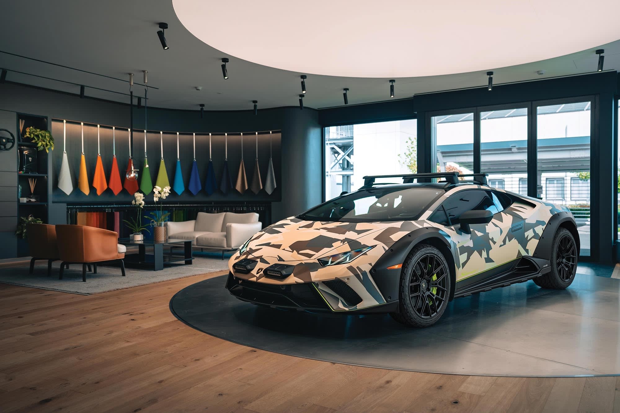 A stylish showroom featuring a camouflaged-painted Lamborghini sports car in the foreground. The room is modern with a wooden floor, and the background showcases a wall with a variety of colorful material samples hanging in a row