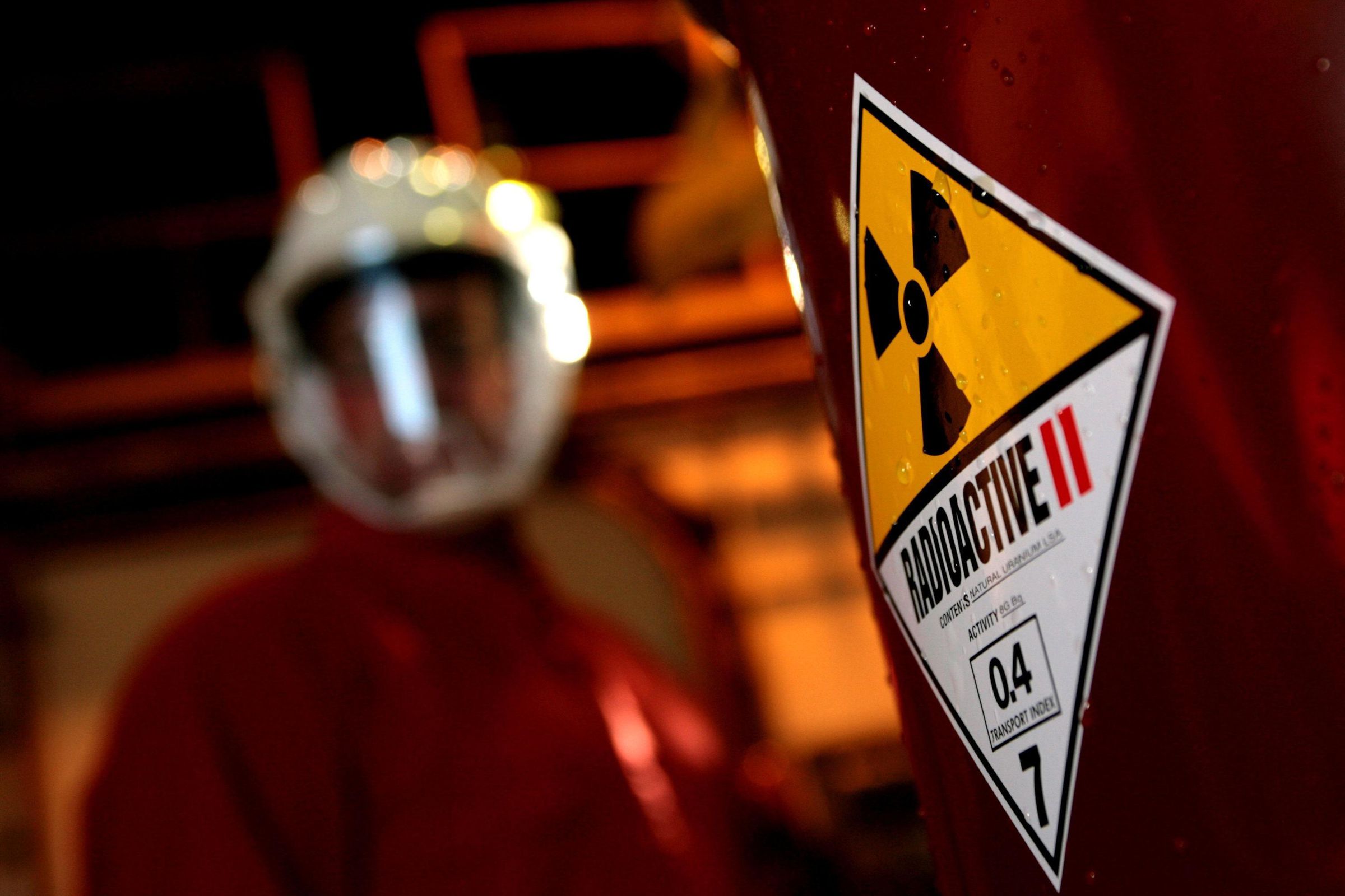 A worker wearing a mask stands behind a drum labeled with a sticker that says “radioactive.”