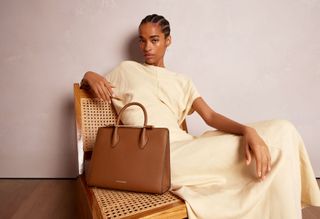 a model wears a cream-colored dress and poses with a brown Strathberry handbag