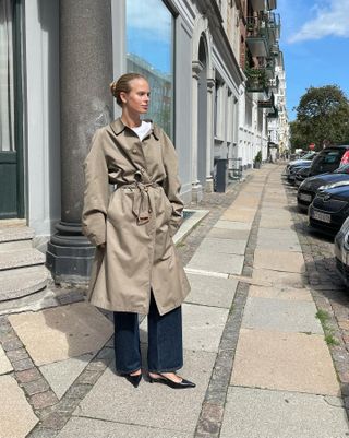 Scandinavian creative and influencer Clara Dyrhauge poses on a sidewalk in Copenhagen with a simple bun, belted trench coat, white t-shirt, dark-wash jeans, and black slingback kitten heels