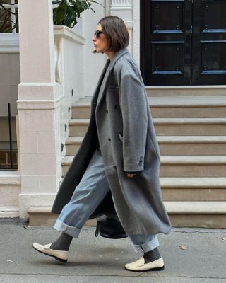 British influencer Lucy Alston walks on London sidewalk gray long coat, cuffed jeans, gray socks, and cream loafers