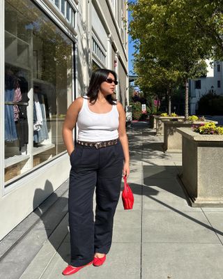 influencer Marina Torres poses on a sidewalk wearing retro oval sunglasses, a white tank top, studded belt, black pants, red mini bag, and classic red ballet flats