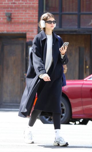 Kaia Gerber wearing headphones, black sunglasses, a long coat, a white top, black leggings, Nike socks, and Nike V2K run sneakers.