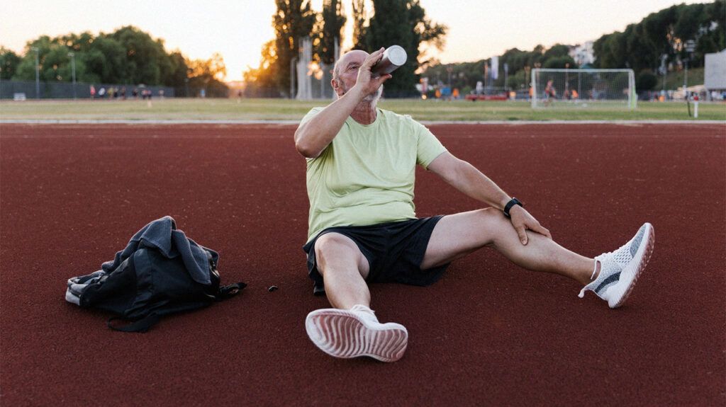 older white man in sports equipment sitting on running track drinking water