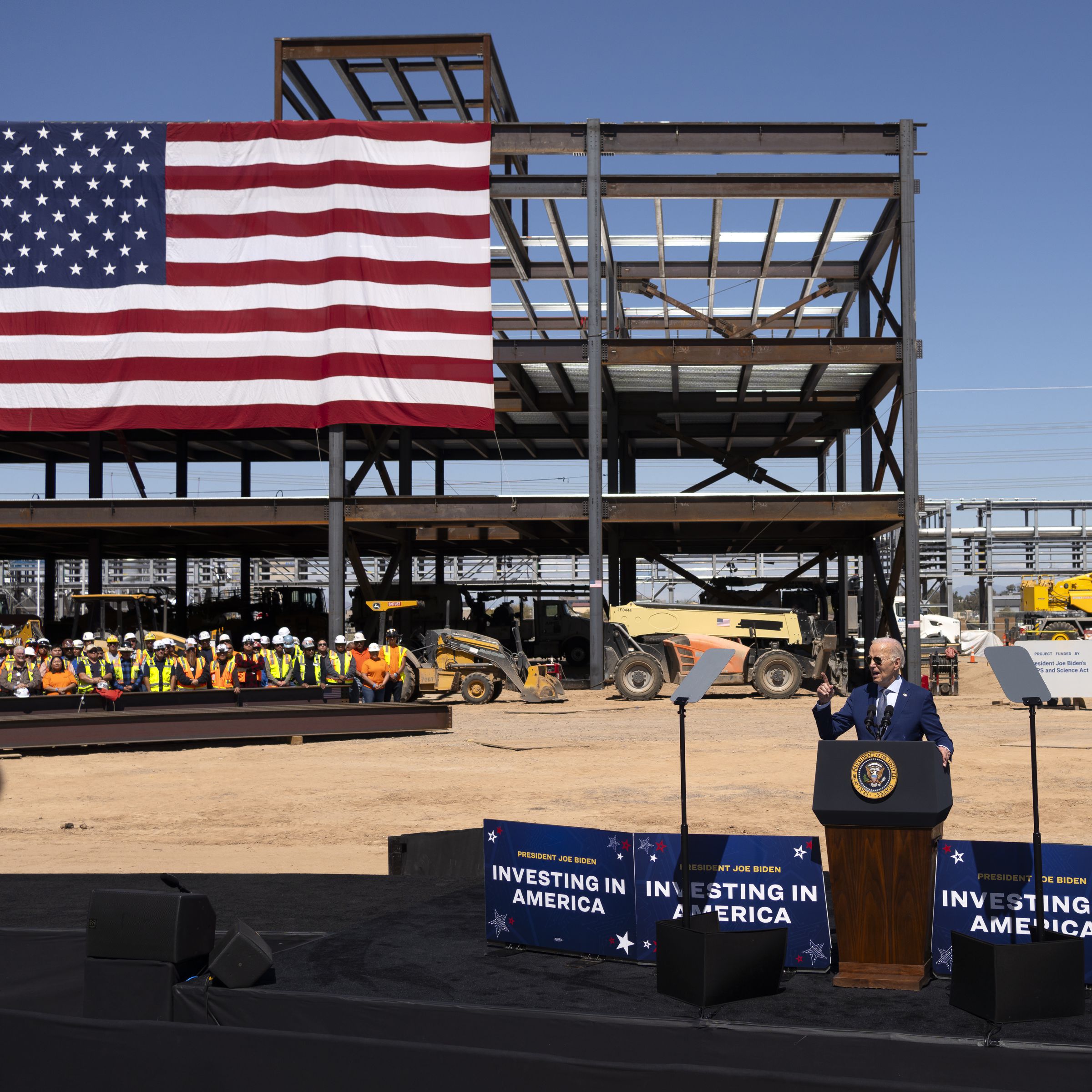 Joe Biden speaks at a podium in the middle of a construction site. Workers in bright vests and hard hats can be seen standing behind him, with a large American flag hung above them. Construction equipment is in the background.