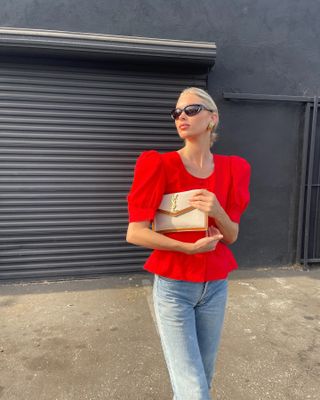 Elsa Hosk wears a red peplum top, light-wash straight-leg jeans, black sunglasses, chunky gold earrings, and a beige Saint Laurent clutch.