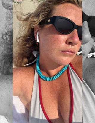 Influencer Martine Lavelle poses at a Cape Cod beach wearing black cat eye Crap sunglasses, AirPods, turquoise beaded necklace, and a gray Frankies Bikinis bikini top with red contrast trim.