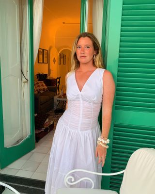 Influencer Martine Lavelle poses on a balcony of a seaside hotel in Italy with bright green shutters wearing a sleeveless white ruched dress and shell embellished bangles.