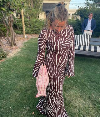 Influencer Martine Lavelle poses in a backyard wearing a half-up hairstyle, cord necklace with pendant, animal print Realisation Gia dress, and fish-shaped bag.