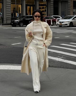 Caroline Lin wears an ivory trench coat, an ivory sheer top, a white belt, white trousers, and black heels.