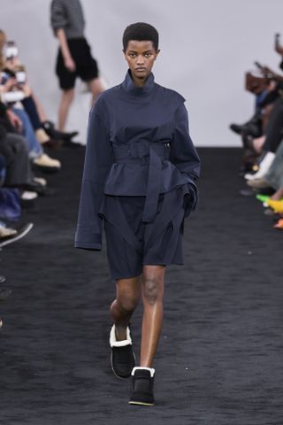 JW Anderson fall/winter 2024 runway shot featuring a model wearing a navy top with a double belt over it, navy shorts, and black shearling shoes.