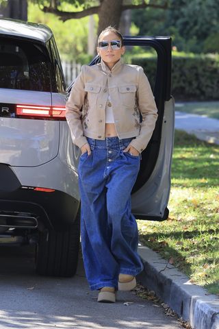 Jennifer Lopez wears pleated wide-leg jeans, platform chestnut boots, a beige denim jacket, cropped white shirt, and large silver sunglasses.