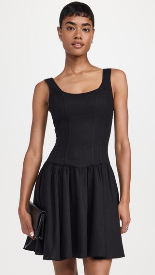 a model wears a short black scoop-neck sleeveless mini dress with a pleated skirt