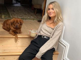 British influencer Lucy Williams poses on steps in her London home with her dog wearing a neutral cardigan and matching top, black belt, and black pants