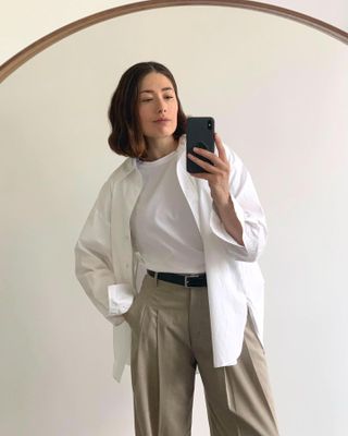 fashion influencer Lucy Alston poses for a mirror selfie with a short bob haircut, a white t-shirt, white button-down shirt, black belt and tan pleated pants