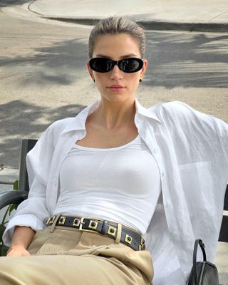 fashion influencer Cass Dimicco poses for a photo while sitting on a bench in Miami wearing black oval sunglasses, a white tank top, open white button-down shirt with pocket, studded balck Aureum belt, tank pants, and a mini black bag from The Row