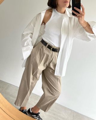 British fashion influencer Lucy Alston poses for a mirror selfie wearing an open white button-front shirt, white ribbed racerback tank top, black belt, cropped pleated khaki pants, and black Adidas Samba sneakers