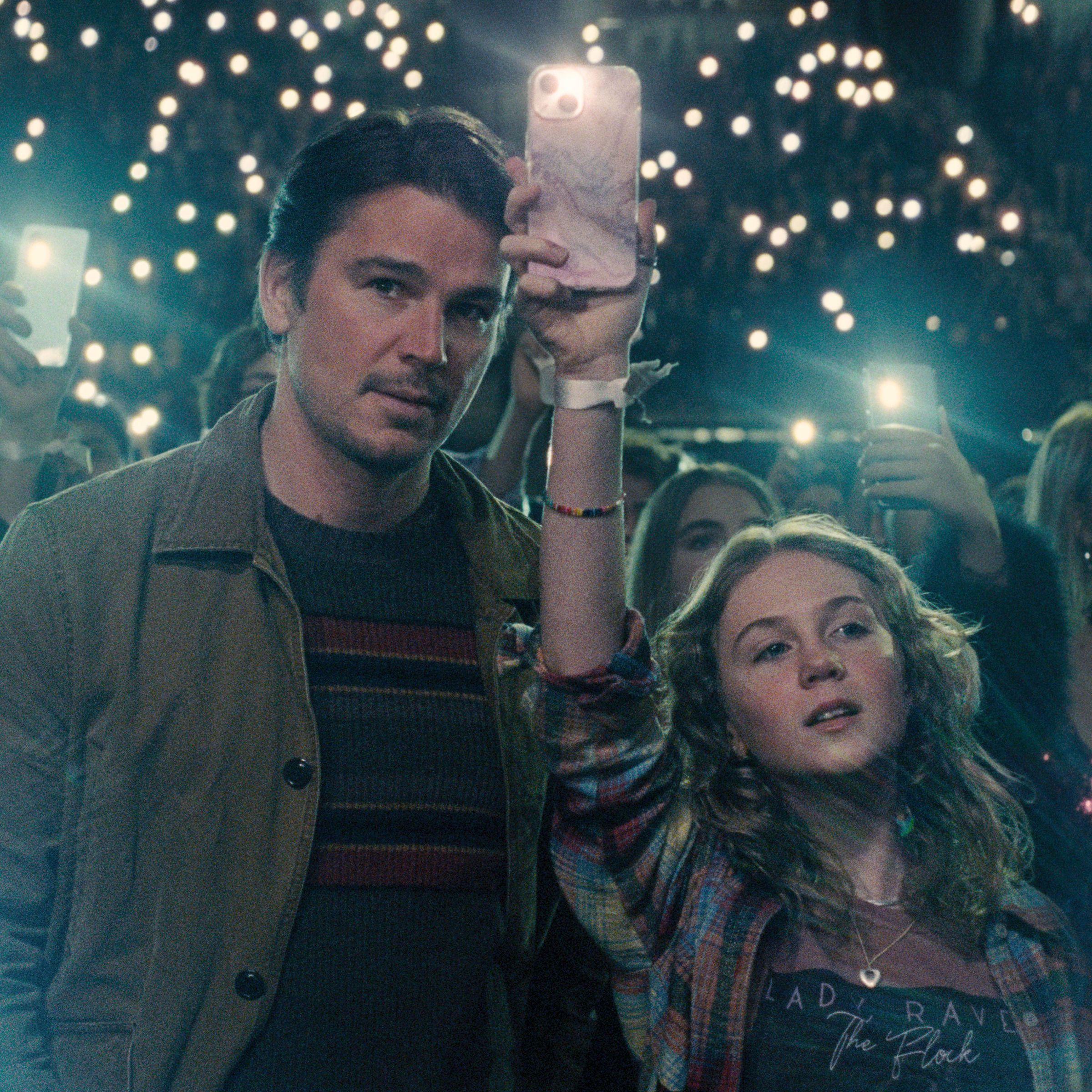 A man and a girl standing next to each other in a concert hall as thousands of people around and behind them hold up their phones.