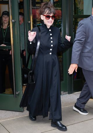 Winona Ryder waves at a crowd wearing a black long coat and black lug-sole boots