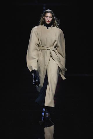 Photo of model wearing oversize camel coat and black gloves, black shoes during Khaite Fall/Winter 2024 runway show.