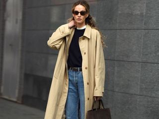 female fashion influencer Anouk Yve poses wearing oval sunglasses, a collared trench coat, a black sweater layered over a white tee, and belted jeans