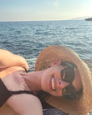 Anne Hathaway wearing black large sunglasses with a straw sun hat and a black swimsuit at the beach.