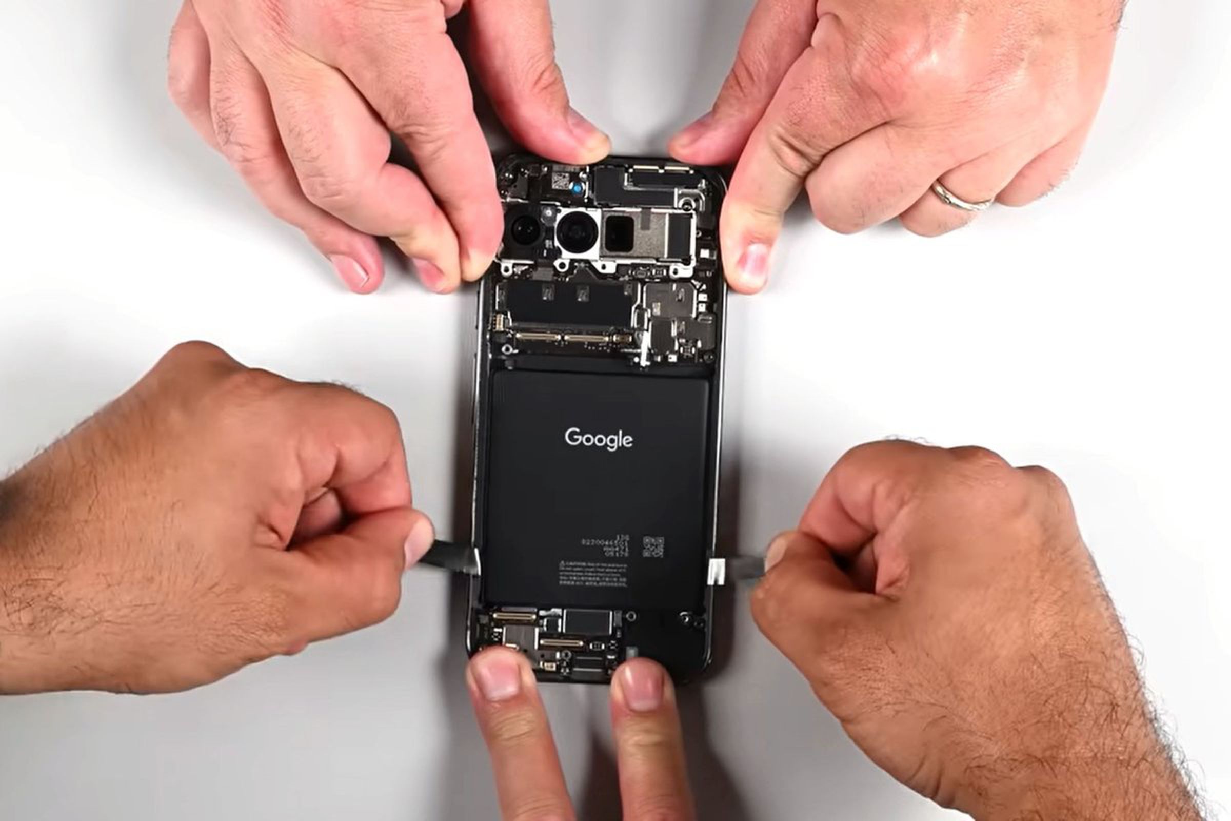 Image of the Pixel 9 Pro Fold internals, with three sets of hands attempting to hold the phone in place and pull out one of the batteries with pull tabs.