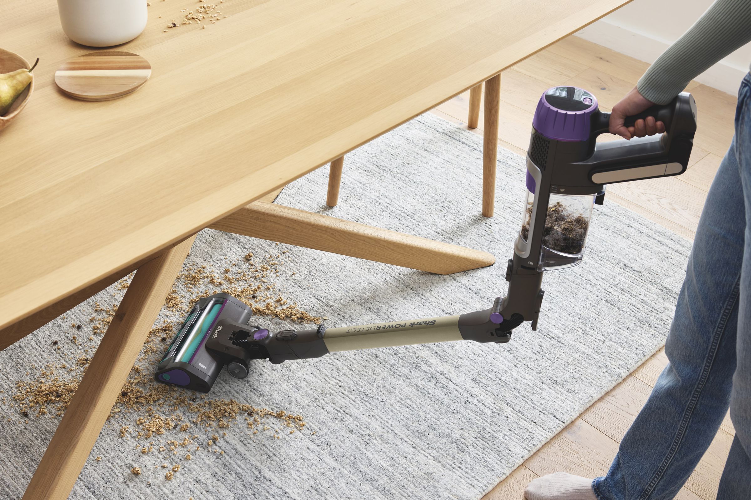 maple table and rug under it with sawdust on top, and purple and silver stick vacuum in person’s hand is folded and vacuuming so person doesn’t have to lean.