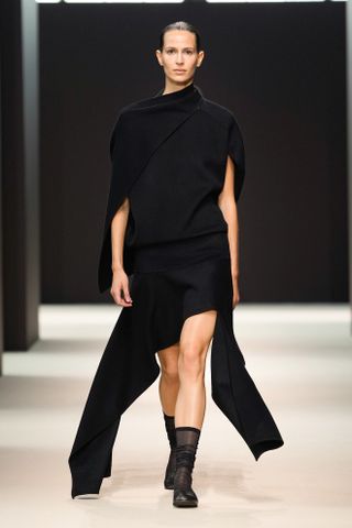 A woman wears a black scarf-like top with an asymmetrical skirt, black sheer socks, and black ballet flats at the Cos F/W 24 show.