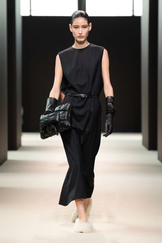 Cos fall 2024 show with a woman wearing a black dress, black belt, padded oversize clutch, and fuzzy shoes.