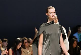 A model at the COS f/w 24 show wearing an army green blouse with a scarf-like neckline.