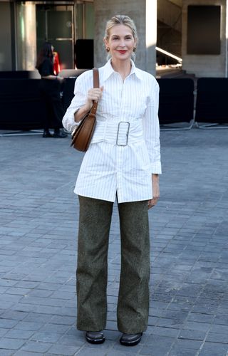 Kelly wears a white pinstripe top, wide-leg pants, black shoes, and a brown shoulder bag.