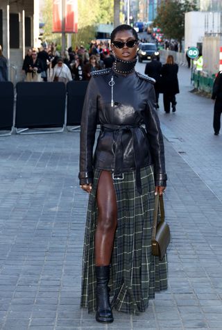 Jodie wears a green check-print skirt, black boots, and a black leather jacket with studs