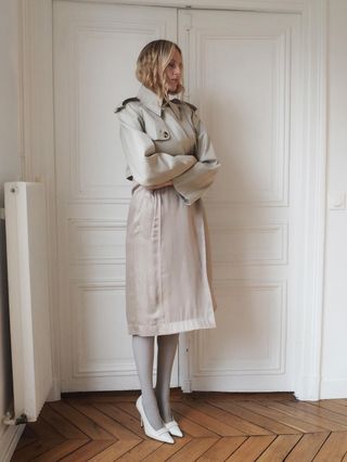 Elly McGaw wearing a taupe trench coat with gray tights and white heels.