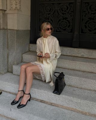 Christie wears an ivory blouse with a tie detail, ivory skirt, black pumps, and a black top-handle bag.