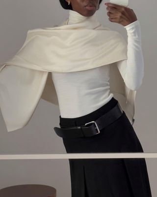 Sylvia wears an ivory cape, a white long-sleeve turtleneck, a black skirt, and black belt.