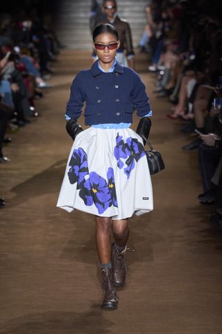 Woman in nayy cardigan and floral skirt on the Miu Miu runway.