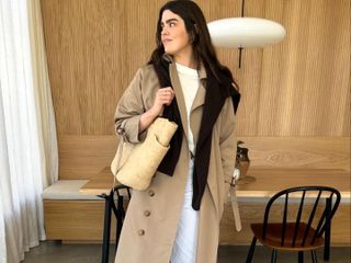 British fashion influencer Anna Howard poses in her modern London kitchen wearing a trench coat, black sweater draped over her shoulders, a white t-shirt, belt, white pants, and a straw tote bag