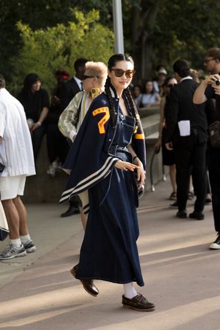 A woman wears a letterman jacket, a denim dress, socks, and boat shoes.