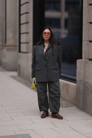 A woman wears a gray blazer, gray trousers, a yellow bag, and brown boat shoes.