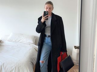 British fashion influencer Brittany Bathgate poses for a mirror selfie wearing a black maxi coat, gray t-shirt, black belt, and ripped distressed jeans