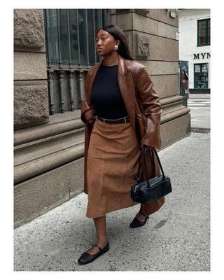 A woman wears a brown leather jacket, black shirt, black belt, suede skirt, black flats, and a black east-west bag.