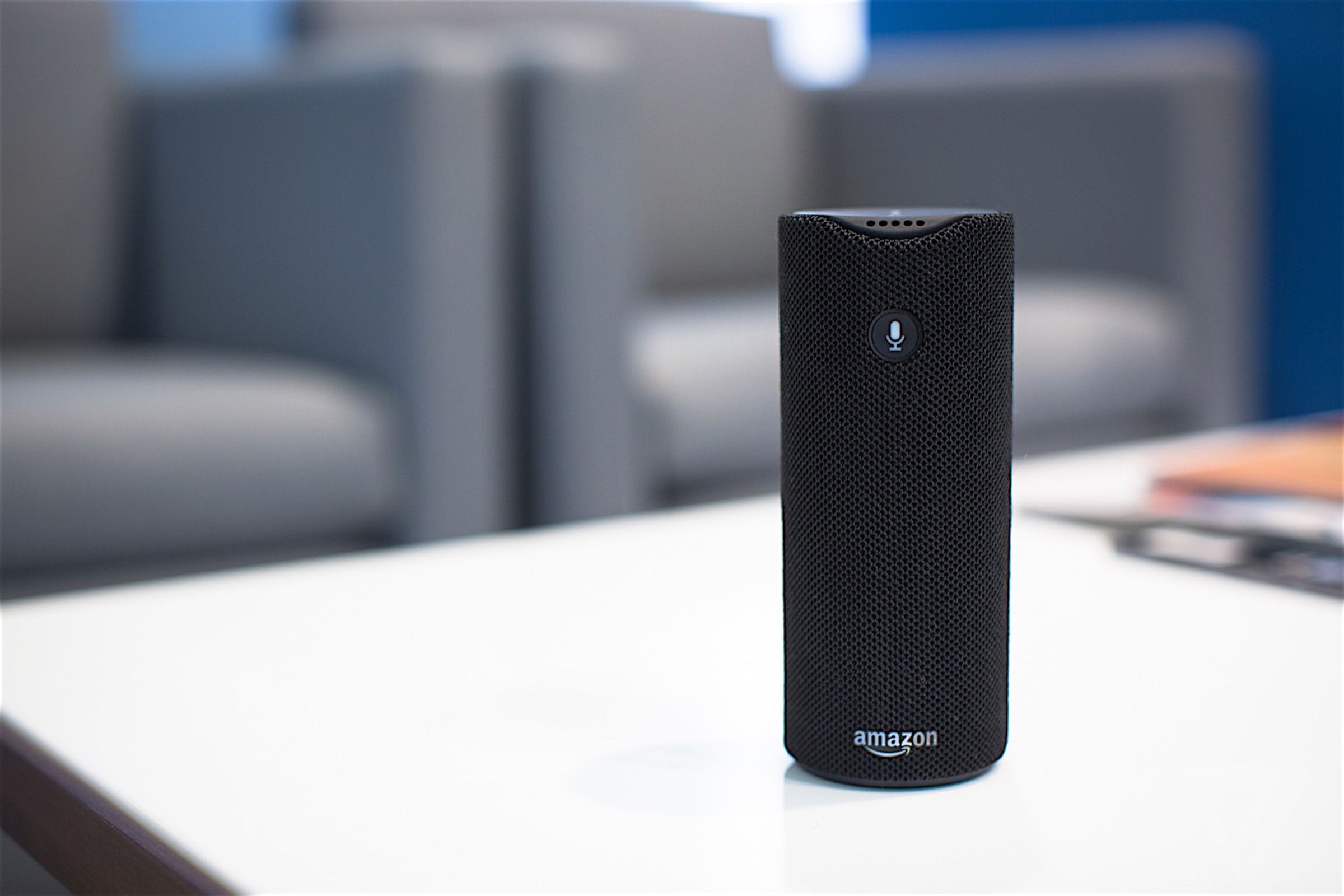 The Amazon Tap smart speaker sitting on a white coffee table in front of a pair of chairs.