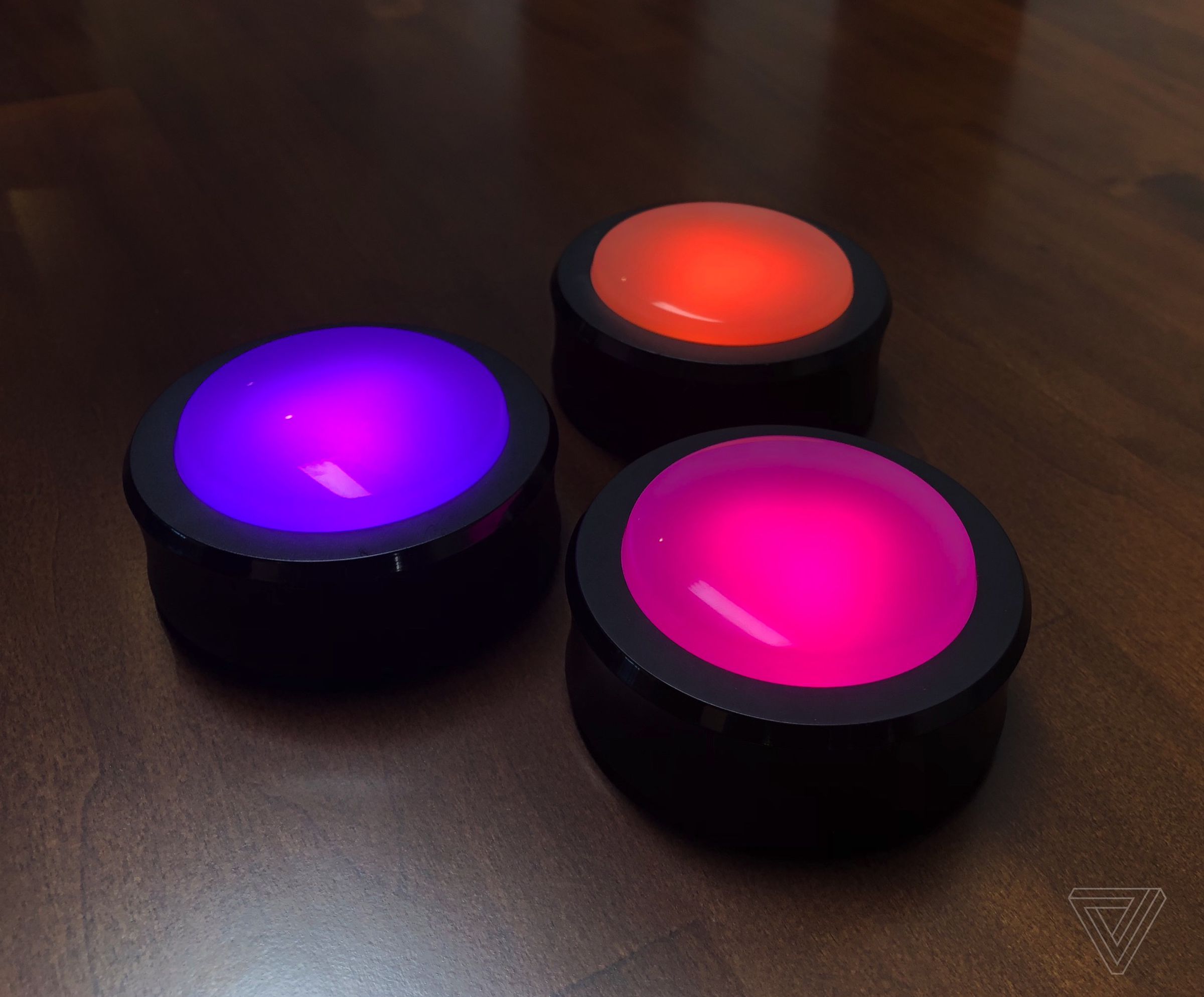 Three Amazon Echo Buttons glowing purple, pink, and orange, on a wooden desk.