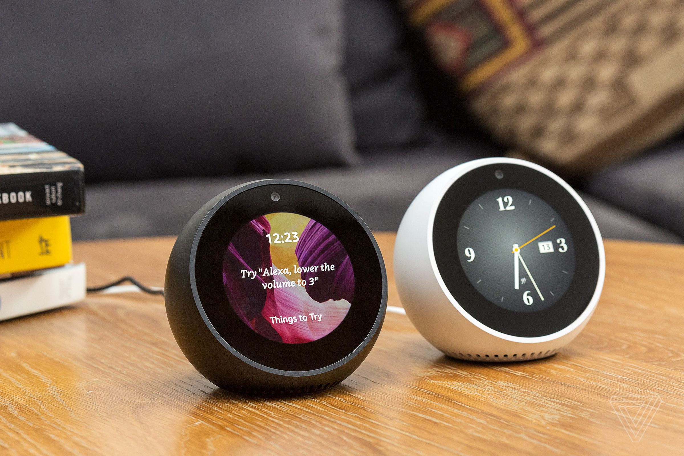 A black and a white Amazon Echo Spot smart speaker sitting side-by-side on a wooden table.