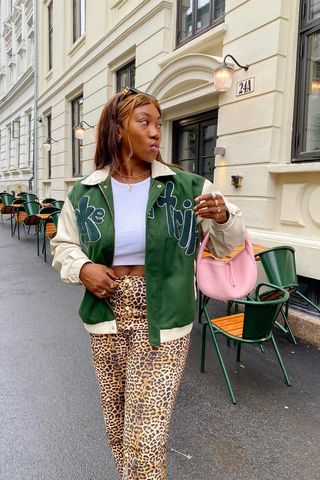 A woman standing on the sidewalk wearing a green bomber jacket over a cropped white tank top, cheetah print pants, and pink crescent bag.