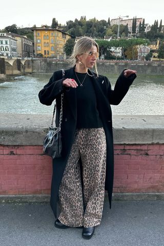 A woman walking down the sidewalk wearing a long black coat with a black crewneck sweater, leopard print pants, and black ankle boots.